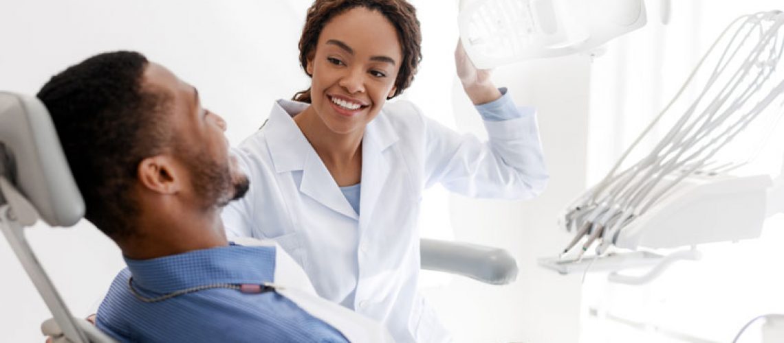 Male patient sitting in dental office, female dentist turning on a lamp before treatment, smiling to male patient.
