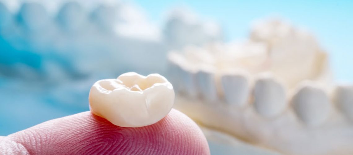 a dentist’s finger holding a dental crown with a dental bridge model behind.